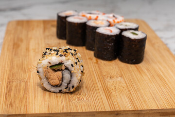 Sushi roll sits on a wooden cutting board. The sushi roll is surrounded by other sushi rolls, creating a sense of abundance and variety