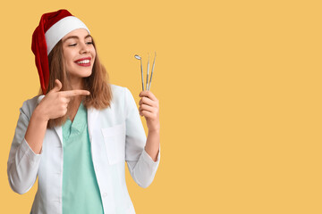 Young female dentist in Santa hat pointing at dental tools on yellow background
