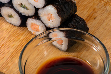 Glass bowl filled with soy sauce sits beside a neatly arranged pyramid of sushi rolls, each featuring a different type of fish. Elements rest on wooden board, creating a warm and inviting composition