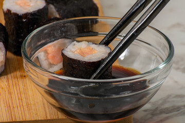 Chopsticks grasp a piece of sushi dipped in soy sauce within a clear glass bowl. In the background, various sushi rolls are arranged on a wooden surface, showcasing Japanese culinary artistry.