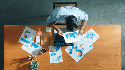 Top aerial view of manager working on laptop while looking at data analysis. Leader compare financial statistic graph while making decision at meeting table with document scatter around. Convocation.