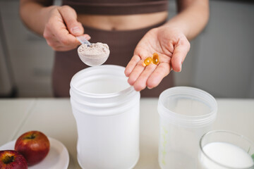 Athletic woman in sportswear preparing protein drink in a kitchen, holding a portion of whey protein powder in a measuring spoon and omega-3 supplement capsules in her hand