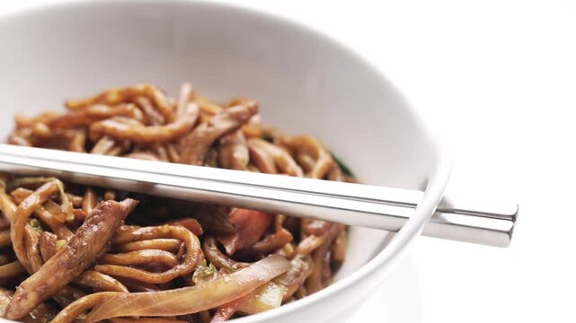 A pair of metal chopsticks resting on a serving of Chinese lo mein noodles with chicken in a white bowl.