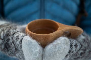 Close-up of hands in knitted mittens holding a wooden cup, with a blue jacket in the background. Concept of warmth and coziness outdoors