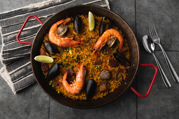Classic Spanish dishes, seafood paella in a traditional skillet against the rustic blue concrete backdrop. Spanish paella with shrimp, hugs, mussels, green peas, and fresh lemon wedges from above.