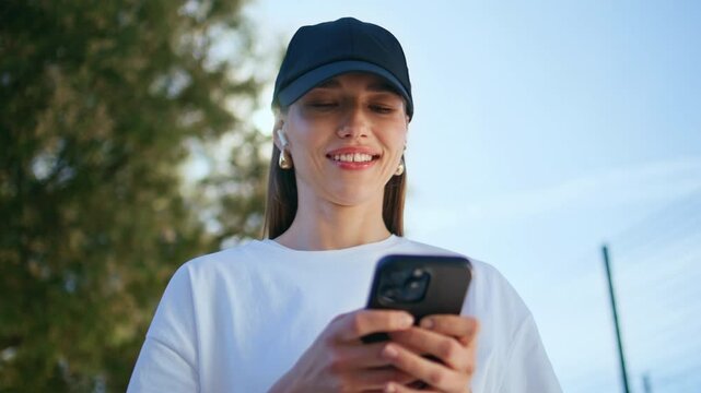 Smiling girl texting smartphone at city street wearing cap closeup. Happy woman