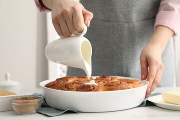 Woman adding frosting onto freshly baked cinnamon rolls at white table indoors, closeup