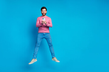 Happy young man with beard jumping joyfully while holding a smartphone against a bright blue...