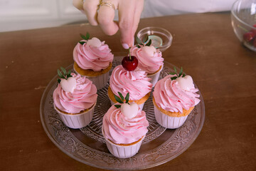 A woman's hand arranges cherries on a vanilla homemade holiday cupcake with whipped cream, on a wooden table, copy space. Holiday, Sunday breakfast and relaxation. Selective focus.