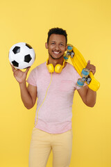 Handsome african american young man wearing headphones holding soccer ball and skateboard over yellow background.