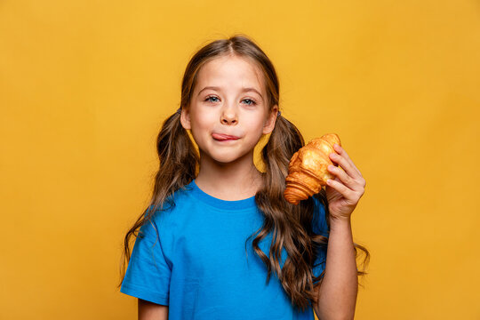Happy smiling little girl in blue t-shirt eating French croissant on yellow background, and licks her lips with pleasure. Copy space. High quality photo. French breakfast concept.