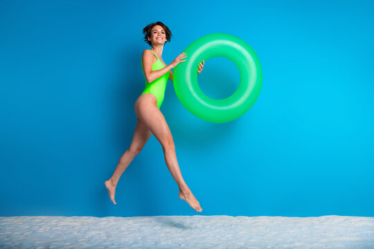 Young woman holding a green inflatable ring in a swimsuit leaping joyfully against a blue background