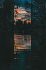 Sunset over a river with trees and between two buildings silhouetted against the glowing sky. Ideal...