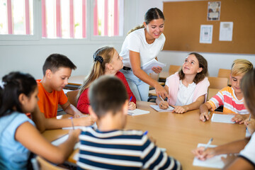 Young female teacher conducting lesson with interested preteen children sitting around common desk in classroom