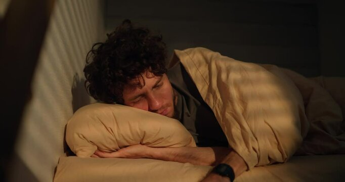 A sleepy man with curly hair wakes up in the morning of his bed and looks at the phone screen at the alarm clock in the morning
