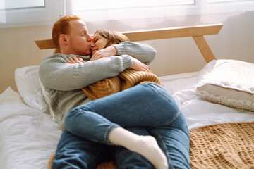 A happy young couple man and woman are hugging each other while lying together on bed at home. Kissing and smiling. Romantic day together.