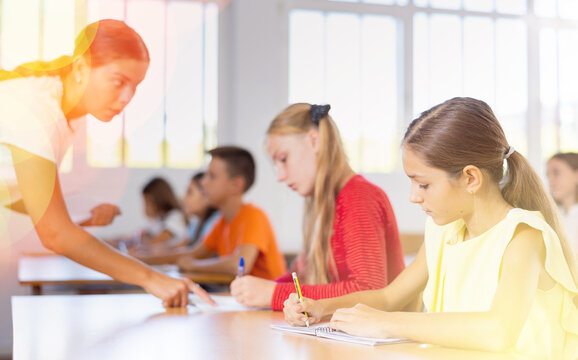 Interested intelligent preteen girl studying in classroom with classmates, listening attentively to teacher and writing in notebook