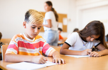 Portrait of concentrated tween boy studying in classroom, listening to schoolteacher and writing in...
