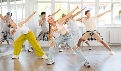 During rehearsal workout, teenage girl dances hip hop with friends against background of large mirror, window. Aerobics students enjoy hip hop break dancing, perform modern dance.