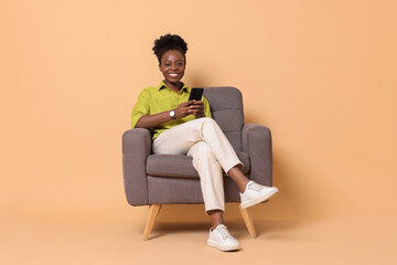 Smiling woman with smartphone sitting in armchair on beige background