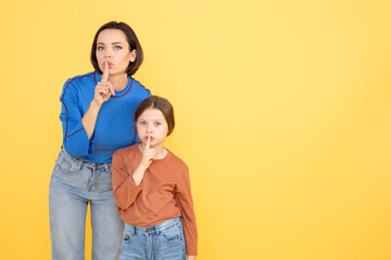 A mother and her daughter stand close together against a bright yellow background, both making a shh gesture with their fingers, copy space