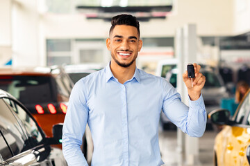 Happy muslim guy atnding among nice luxury and sports cars in huge auto showroom, showing key from...