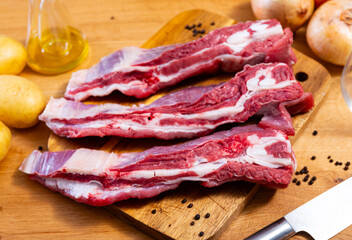 Table is laid out with products for cooking lunch, baked meat and side dishes. Raw beef ribs on board, potato tubers and whole onions, small carafe with olive oil are necessary ingredients for dish
