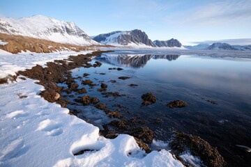 Obraz premium A breathtaking winter landscape features serene snow-covered terrain and still water reflecting majestic mountains under a clear blue sky, creating a scene of tranquility.