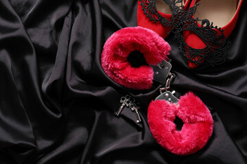Red fluffy handcuffs, keys, shoes and lace mask on black fabric, flat lay. Space for text