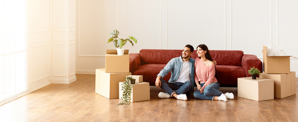 Moving Day. Lovely couple sitting on the floor in new home with cardboard boxes around. Happy man...