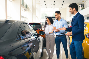 Cheerful middle eastern couple looking at black sports car, choosing new auto at newest automobile dealership, having conversation with male sales associate , full length shot, copy space