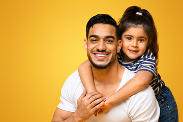 Obraz premium Portrait Of Happy Young Middle Eastern Dad And Cute Little Daughter Posing Together Over Yellow Background, Female Kid Embracing Daddy From The Back And Smiling At Camera, Panorama, Copy Space