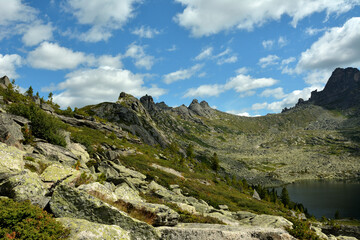 Rocky gentle slopes of a mountain range with bushes and cedars under a summer cloudy sky.