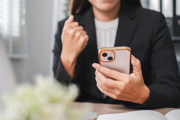 A professional woman in a suit celebrates a successful moment while looking at her smartphone in a contemporary office environment.