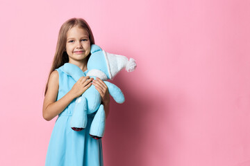 Adorable little girl in a blue dress holding a teddy bear on a vibrant pink background, radiating...