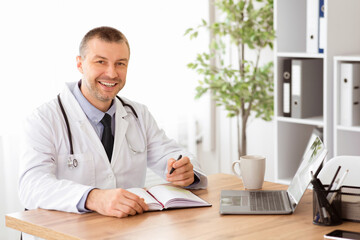 Fototapeta premium Professional Help Concept. Portrait of happy handsome male doctor in stethoscope and white unifrom sitting at table with laptop at his office, posing, looking at camera and writing in his notebook