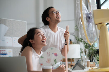 Two young Asian startup coworkers are fun blowing wind with electric fan, working in hot...