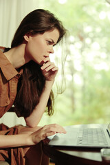 A focused young woman in a brown dress working on a laptop, deep in thought against a green nature...