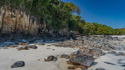 Picturesque boulders are scattered on the sandy beach. Stones on the white sand.  Green vegetation on the coastal cliffs. Clear blue sky. Madagascar. Nosy Be 