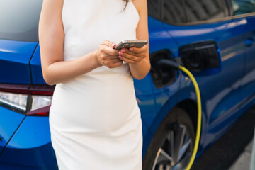 Woman charging a blue electric car while using smartphone in a sunny day