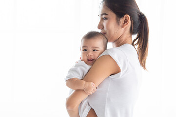 Asian beautiful young single mother holding smiling newborn baby in bright indoor. Both dressed in white, sharing joyful and affectionate moment home environment. Baby healthcare products concept