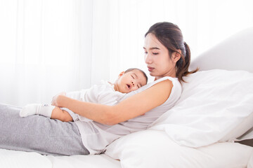 Asian young beautiful mother resting on white bed with sleeping newborn baby on her chest. Both dressed in soft white clothing, tired women sleep from taking care adorable infant with love and tender