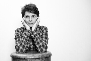 Nachdenkliche Person in kariertem Hemd stützt Ellenbogen auf Hocker in Schwarz-Weiß-Fotografie, vor hellen Hintergrund