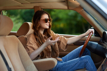 Young woman in stylish sunglasses joyfully talking while driving a vintage car, showcasing casual fashion in a vibrant green setting.