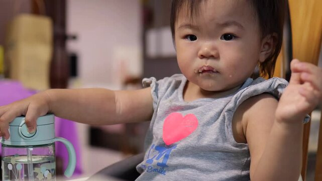 Baby girl enjoys her mealtime, holding a food container and sippy cup while sitting on a high chair