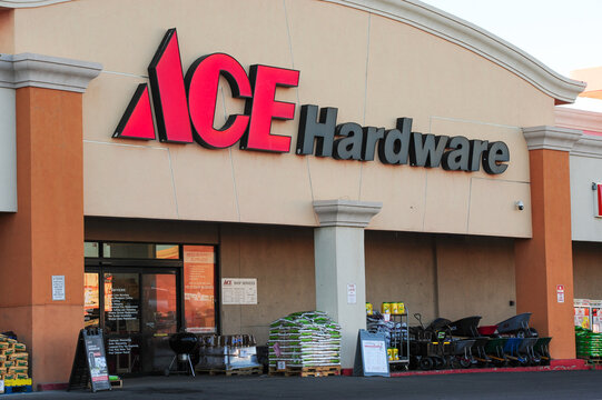 The storefront of an Ace Hardware store featuring a prominent red and black logo, products like wheelbarrows, garden supplies, and bags of fertilizer displayed outside the store