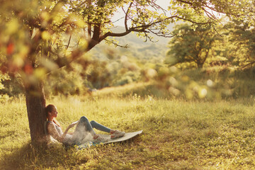 A young woman relaxing under a tree, enjoying a book in a serene green landscape during golden...