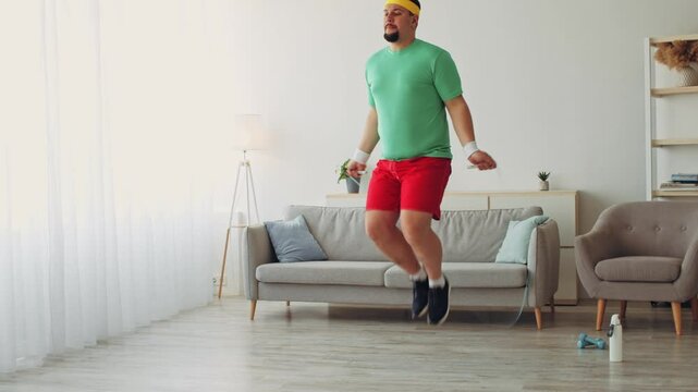 A chubby young man in a green shirt and red shorts jumps rope in his modern living room. He's focused on his fitness routine and seeks to improve his health through regular exercise.
