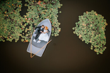 Aerial view of a couple in wedding attire lying in a small rowboat on a pond surrounded by lily...