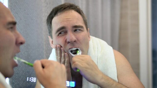 The guy in the reflection of the mirror is brushing his white teeth. Young man with toothbrush suffering from toothache in bathroom